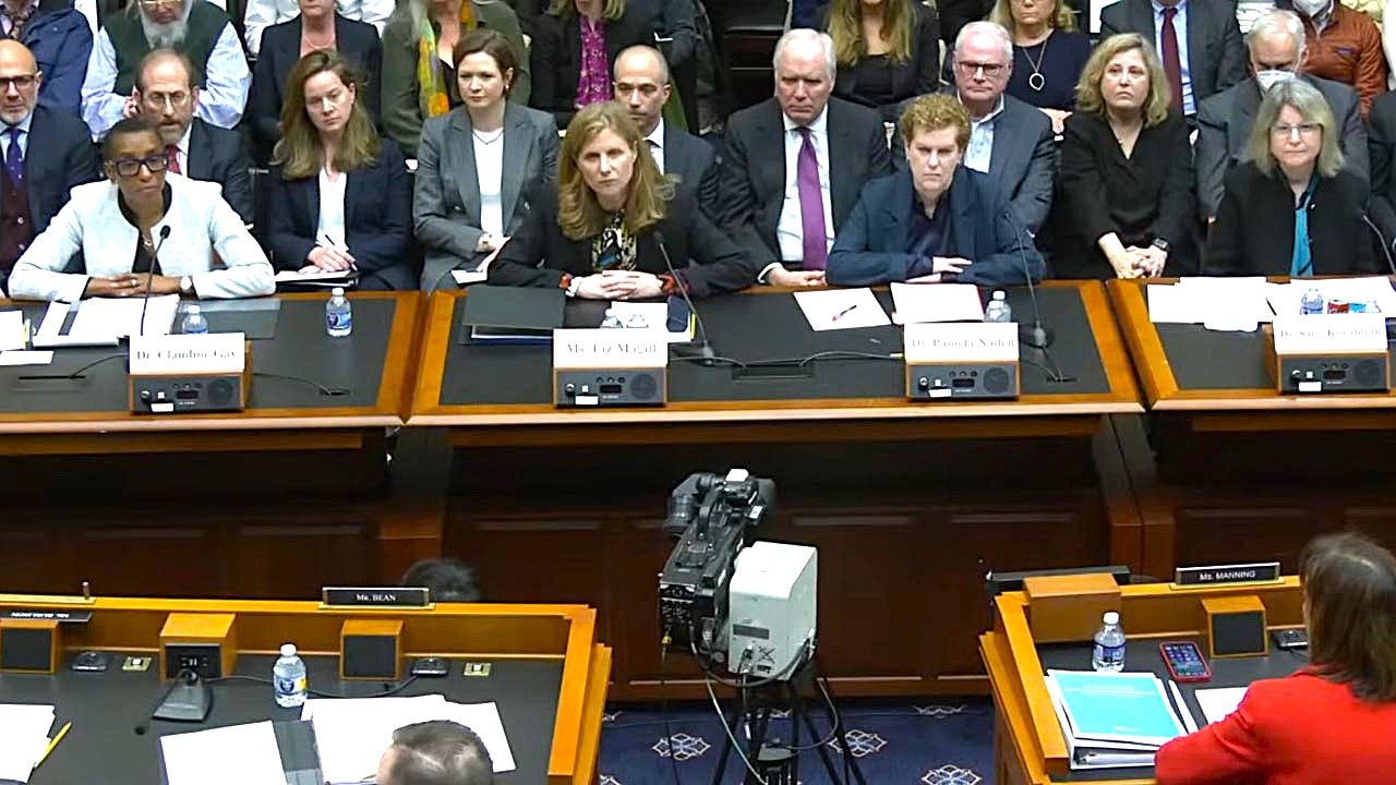 With rows of spectators behind them, four women with serious expressions on their faces sit behind microphones.